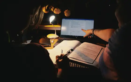 Uczeń przy biurku nocą z lampką, laptopem i zeszytem